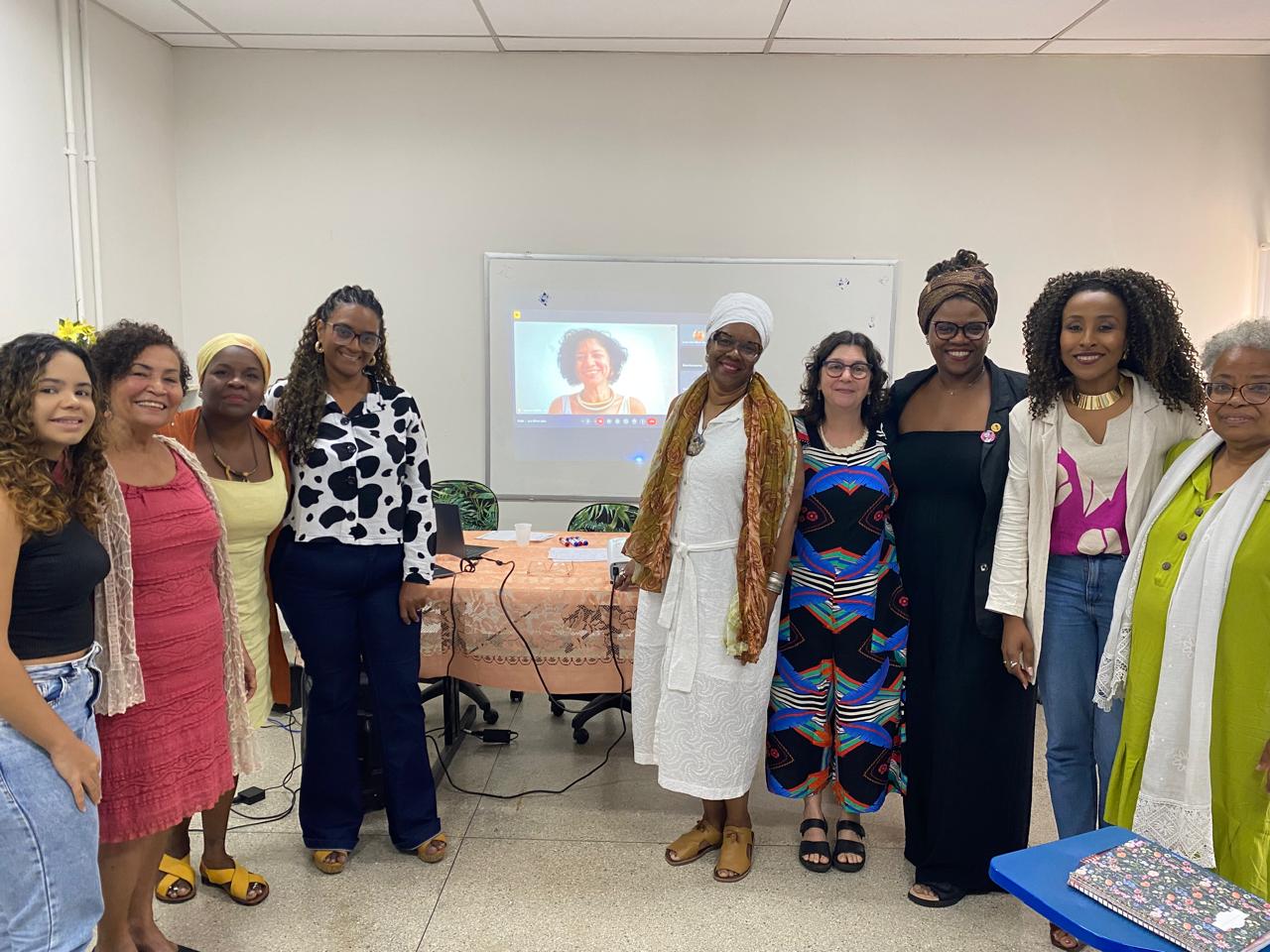 Foto. Área interna. 8 mulheres negras e 1 branca de diferentes idades e estilos de vestimenta. Ao fundo uma projeção mostrando uma video chamada com outra mulher negra.