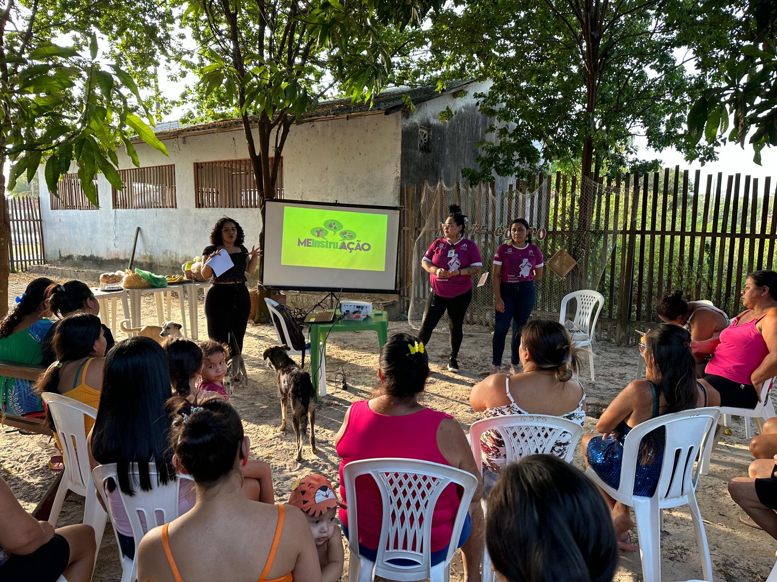 Foto. Área externa. 3 mulheres fazem uma apresentação com projetor. Diversas mulheres sentadas em semicírculo em cadeiras plásticas assistindo