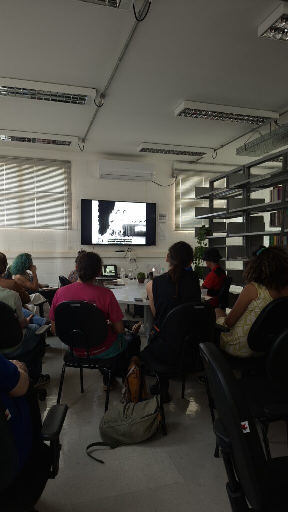 A imagem mostra alunos no curso olhando para o filme que está sendo exibido no curso de Agustin. Estão na biblioteca do Pagu.